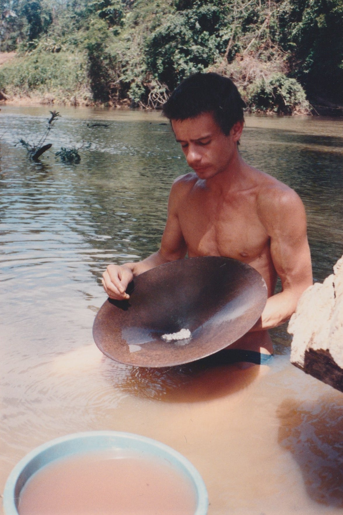 Man holding a pan with gold collected in the centre while standing in a river with trees in the background