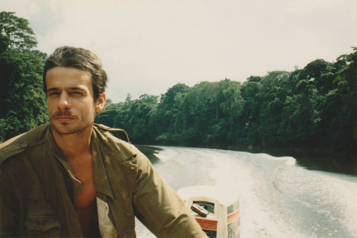 Man in a green jacket standing on a riverbank with dense forest in the background